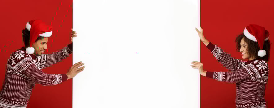 Winter, confrontation and Xmas mood. Cheerful young african american couple in Xmas hats and sweaters, push white billboard with empty space for advert isolated on red background, panorama, side view photo