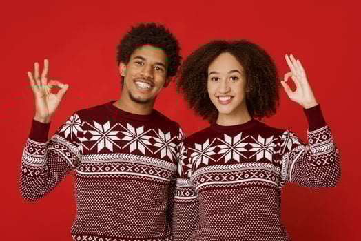 Great New Year and Christmas party. Joyful african american family in identical sweaters show sign okay with their hands and look at camera, isolated on red background, empty space, studio shot photo
