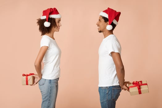 Romantic Christmas for two. Smiling young man and woman in Santa Claus hats look at each other and hold surprise gifts behind backs isolated on light background, profile, studio shot, copy space photo