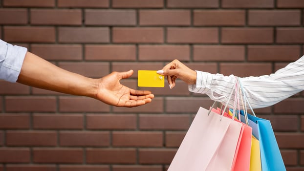 Shopping in boutique. African american girl with multicolored packages gives credit card to seller on brick wall background, cropped, free space photo