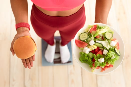 Weight Loss Diet. Fit African Girl Choosing Between Salad And Burger Standing On Scales Indoors. High-Angle, Cropped photo