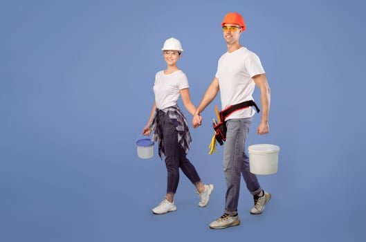 Smiling couple of painters in hard hats and protective glasses holding hands walking with paint buckets over blue studio background, ready for renovation. Full length shot of coating workers family photo