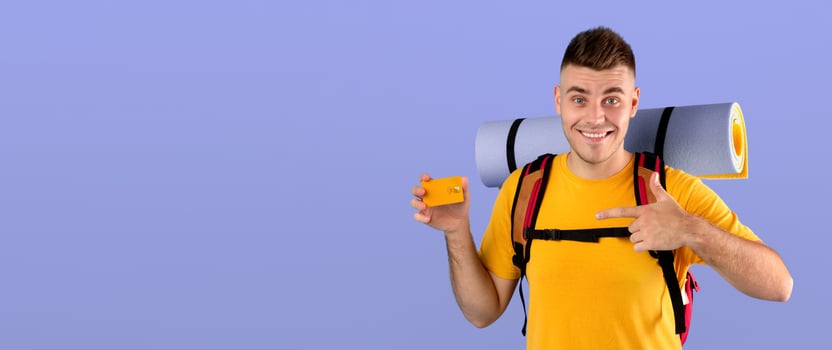 Travel budget concept. Handsome young guy with tourist equipment pointing at credit card over blue studio background. Cheerful millennial man planning funds for his camping adventure photo