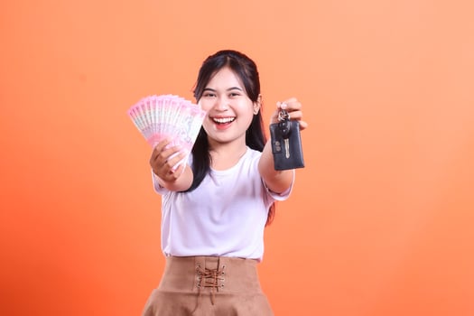Delighted young Asian woman showcasing a fan of money and car keys with a cheerful smile. isolated on orange background. photo