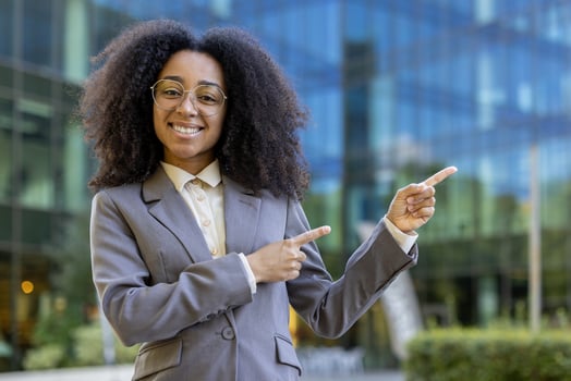 Confident African American business woman in formal attire pointing with both hands, smiling warmly in office setting. Depicts success, confidence, positivity, leadership in professional environment photo