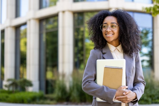 Confident young teacher with glasses stands outside office building holding notebook and tablet. Smiling, professional, and relaxed in natural lighting, conveying success and positivity. photo