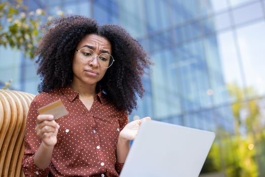 A young woman with curly hair looks puzzled while holding a credit card and using a laptop outdoors near a modern building. photo