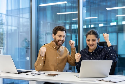 A man and a woman, thrilled by success, celebrate while working together at a modern, bright office space on their laptops. photo