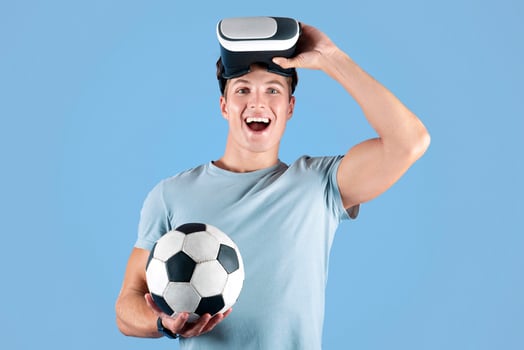 Exercising with virtual reality concept. Young athletic guy in VR headset holding soccer ball on lilac studio background. Positive young sportsman using modern technologies for football workout photo