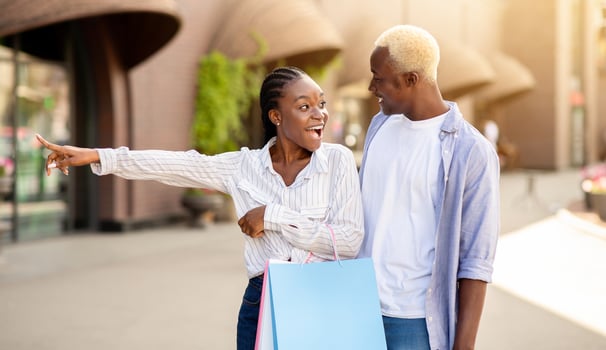 Brand labels and special discounts. Surprised african american girl with shopping bags shows on showcase to guy, on street photo