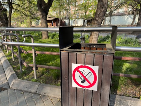Outdoor wooden trash bin with no smoking sign, located in park area with metal railings and green grass on sunny day. Clean environment and public safety concept photo