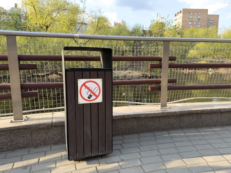 Outdoor trash bin with a clear no smoking sign, promoting public cleanliness and smoke free zones in urban parks or pedestrian areas near water and residential buildings photo