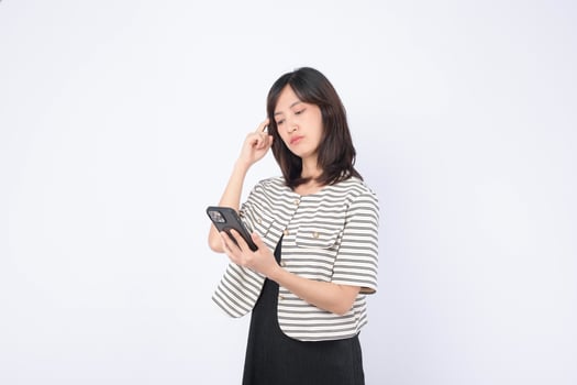 Southeast asian woman is using mobile phone to browse internet with thoughtful expression while standing against plain white background in casual clothing photo