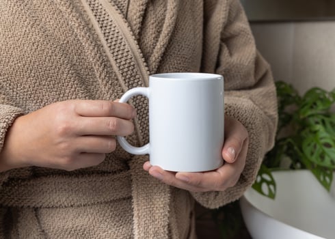 Woman in brown bathrobe holding white blank coffee mug against basin and plant, mockup photo