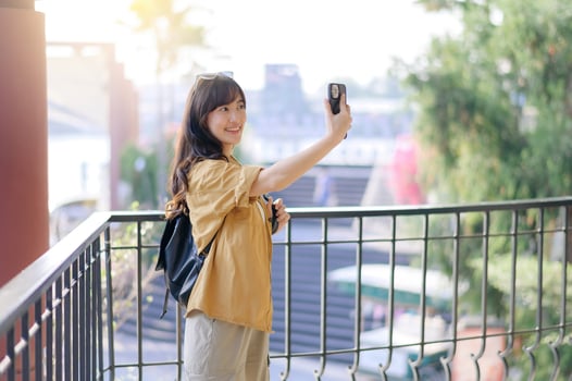 Southeast asian woman travel concept shows happy woman taking selfie outdoors with smartphone wearing casual clothes and backpack in bright sunny day photo