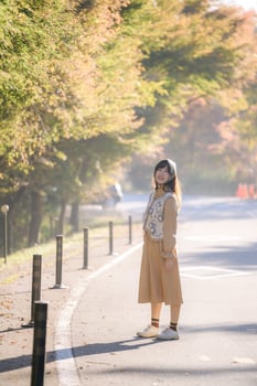 Southeast asian woman stands happily on sunlit road surrounded by autumn trees wearing beige dress and patterned vest enjoying peaceful outdoor scenery photo