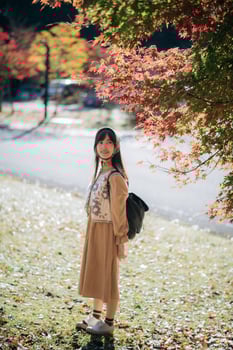 Southeast asian woman enjoys travel concept in autumn park wearing brown dress and backpack under colorful leaves with peaceful and happy expression on sunny day photo