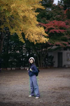 Southeast asian woman wearing hat and casual clothes stands smiling under yellow autumn leaves with backpack in peaceful outdoor setting during fall season photo