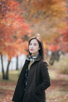 Southeast asian woman stands confidently in dark coat with autumn leaves in background creating warm and peaceful outdoor scene filled with natural beauty and calm expression photo