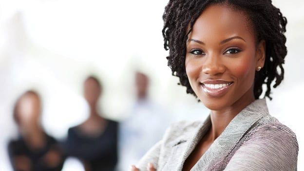 A confident, smiling African American businesswoman with curly hair, wearing a gray blazer, standing in front of a blurred background with other businesspeople. photo