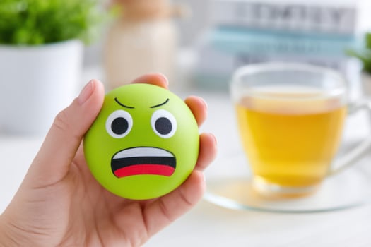 Hand holding a green stress ball with a frowning face, set against a blurred background of a cup of tea and soft greenery, conveying emotions and stress relief photo