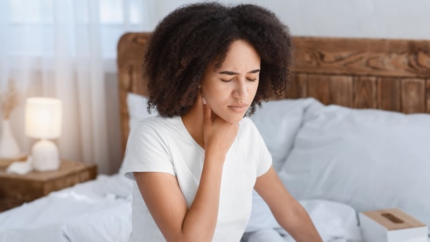 Virus, epidemic, flu, cold, feel bad and health care. Sad young african american lady sits on white bed, suffering from pain and presses hand to throat in interior of cozy comfort bedroom, free space photo