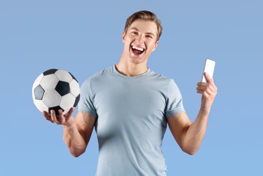 Positive young football fan watching championship online on smartphone, happy over victory of his team on lilac studio background. Millennial guy with soccer ball betting on sports photo