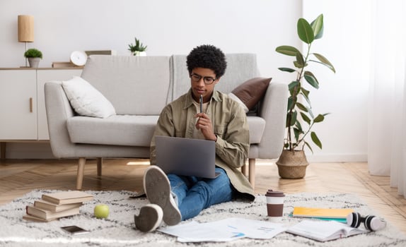 Focused black teen guy sitting on floor with study materials and laptop, at home. African American teenager studying remotely, attending online lecture or seminar, communicating with college teacher photo