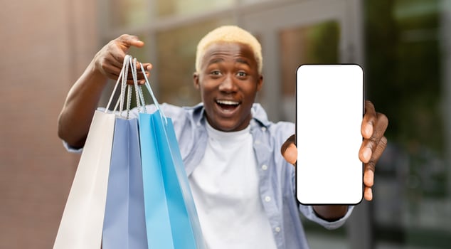 Unbelievable bonus for mobile user, online shopping application. Surprised african american man with packages shows smartphone with blank screen to camera photo