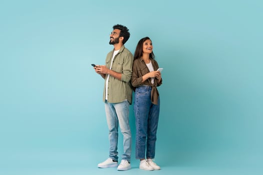 Dating mobile app concept. Stylish young eastern man and woman hipsters standing back to back, using smartphones on colorful studio background, chatting online, looking at copy space, full length photo