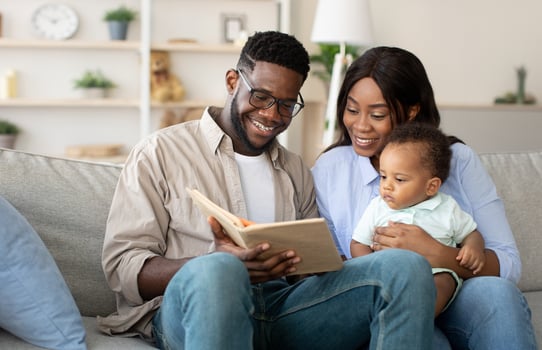 Leisure With Child. Caring Young Parents Reading Book To Their Adorable Infant Son Relaxing Together Sitting On Couch In Living Room, Happy Dad And Mum Bonding With Toddler Baby Kid At Home photo