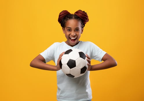 Cheerful afro-american teen girl holding soccer ball and smiling on yellow studio background, happy athletic black female kid playing football. Sport, entertainment, hobby for kids concept photo