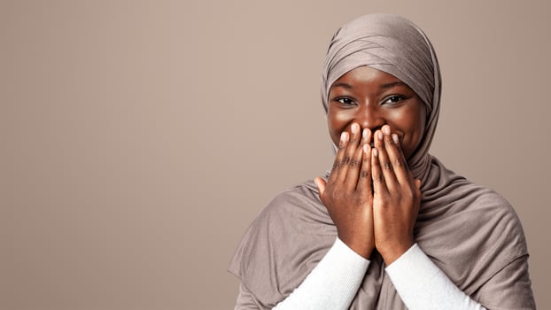 Laughter. Positive Black Muslim Woman In Hijab Covering Mouth With Hands, Feeling Happy Excitement, Emotional Religious Lady Reacting To Pleasant Surprize, Standing Over Light Background, Copy Space photo