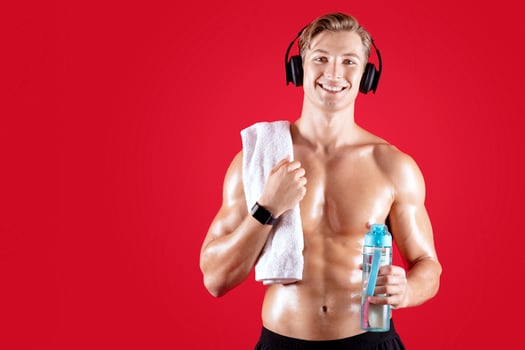 Sexy millennial athlete with bare torso and towel listening to music and holding bottle of water on red studio background. Muscular young guy in headphones keeping hydrated after sauna or workout photo