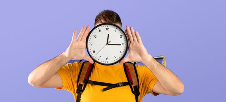 Time to travel concept. Young man with camping equipment holding clock in front of his face over blue studio background. Millennial guy ready to start his hiking journey or vacation photo