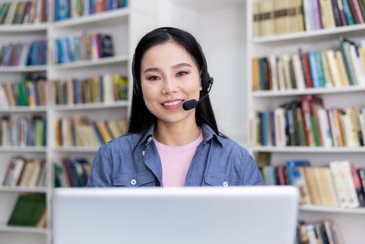 An attractive asian woman smiles while wearing a headset, working or studying remotely. The background shows bookshelves. photo