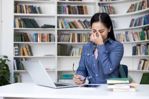 An asian woman rubs her tired eyes while studying at a library. She sits at a desk with a laptop, glasses, and books. photo