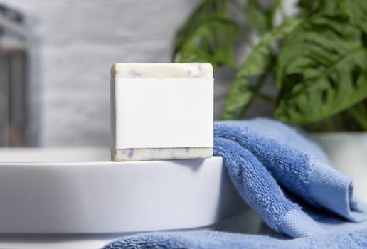 Soap bar with blank label on sink near blue towels and green plant close up, mockup photo