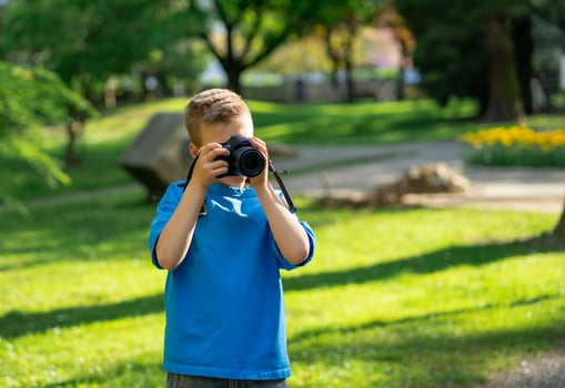 View of cute boy in blue t-shirt taking pictures with camera in city park photo
