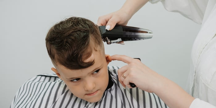 In the picture taken during the hair cutting process by a professional woman holding a special hair clipper in her hands, the child has dark hair. photo