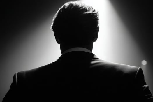 A man stands with his back to the viewer, dressed in a tuxedo. He is illuminated by a single beam of light against a dark background, creating a dramatic atmosphere. The moment conveys anticipation. photo