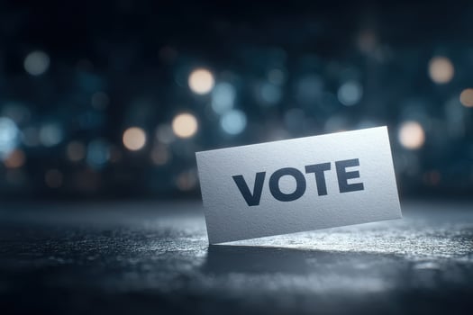 A clear vote sign stands upright on a textured surface, with a blurred background featuring soft lights. The setting captures a moment encouraging civic participation, ideal for promoting elections. photo