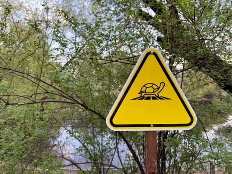 Minimalist turtle crossing warning sign with bold black icon on bright yellow background in natural green park area near water, promoting wildlife safety and environmental awareness without any text photo