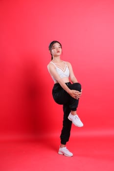 A woman with black pants and a white top stretches her leg, with her hands near her knee, isolated on a red background. photo