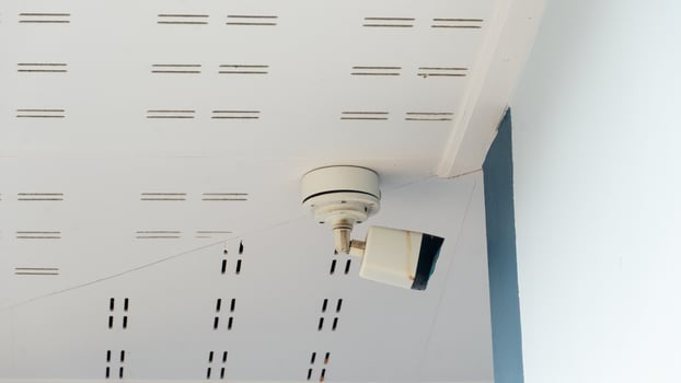 Outdoor CCTV camera. Installed under white ceiling panels. Camera looks towards the side of the building. Material is stained with what looks like rust. photo