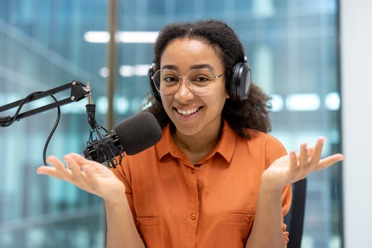 Happy woman hosting a podcast, wearing headphones, gesticulating near a microphone in a modern studio. She's recording a session photo