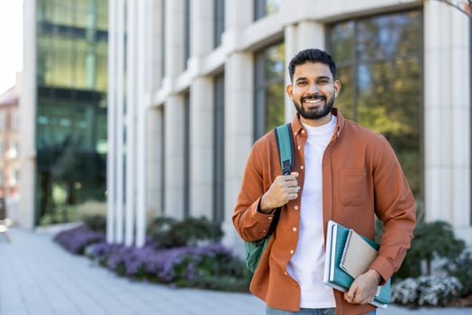 A smiling student poses outside a modern building, holding books and a backpack, ready for classes at a university campus. The scene depicts a beautiful sunny day. photo