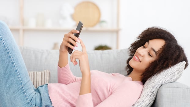 New online game, modern app, dating website, home blog and search information. Happy young pretty african american lady uses smartphone, lies on comfortable sofa in living room interior, free space photo