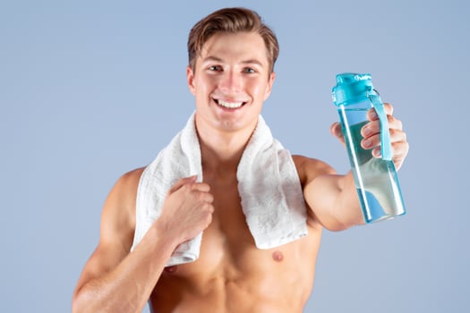 Smiling young sportsman with bare chest and towel showing bottle with fresh water on lilac studio background. Happy fit guy keeping hydrated during sports workout or sauna photo