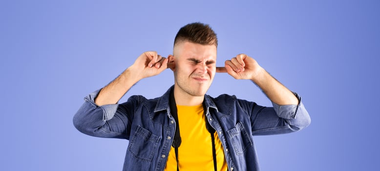 Irritated male traveler with photo camera covering his ears with fingers on blue studio background. Male passenger traveling by plane, suffering from noise during his flight